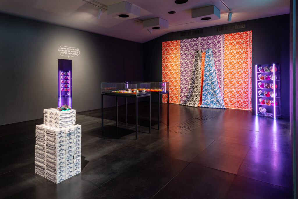 Museo del Novecento in Florence 9 xhibition room of Marion Baruch – Un passo avanti tanti dietro at Museo Novecento, Florence, featuring colorful textile panels on the wall, display cases with objects, stacked boxes, and neon-lit shelves. Photo Leonardo Morfini.