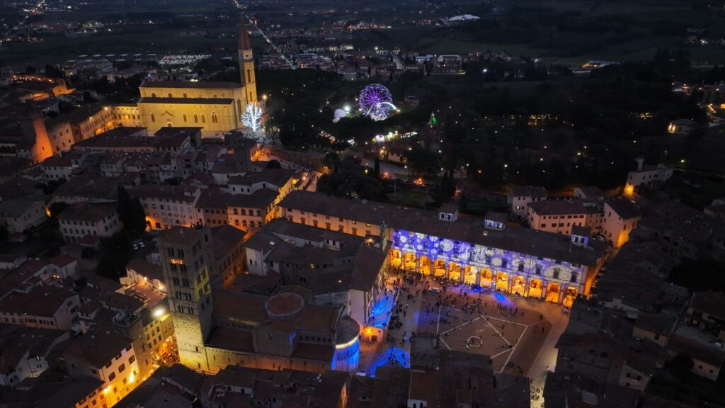Night view of Arezzo showing Piazza Grande lit up and Christmas attractions such as the Ferris wheel and light projections during Arezzo Città del Natale 2025.