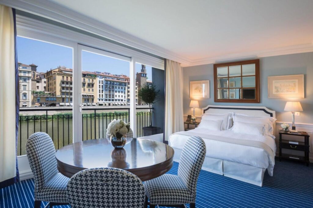 The 7 Best Luxury Hotels in Florence 20 Luxury suite at Hotel Lungarno in Florence with dining table, white bed, and large windows facing the Arno River.