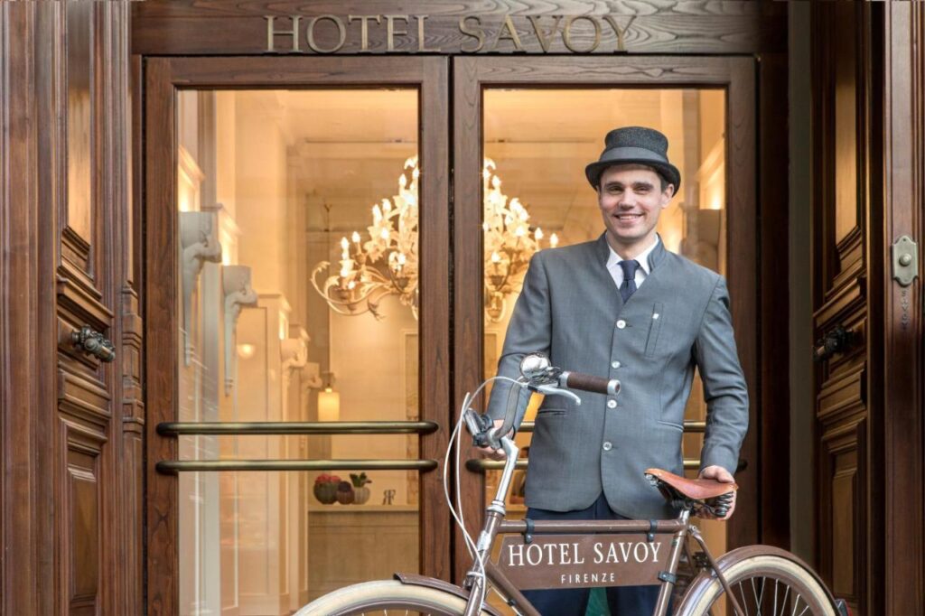 The 7 Best Luxury Hotels in Florence 10 Smiling doorman in vintage-style uniform with a bicycle in front of the entrance to Hotel Savoy Florence, under a wooden sign.