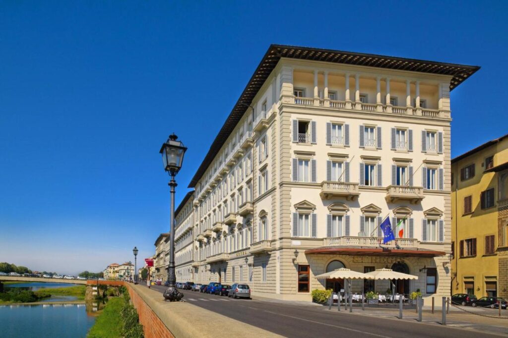 The 7 Best Luxury Hotels in Florence 2 Exterior view of The St. Regis Florence, a grand historic building facing the Arno River under a clear blue sky, with Italian and European Union flags at the entrance.
