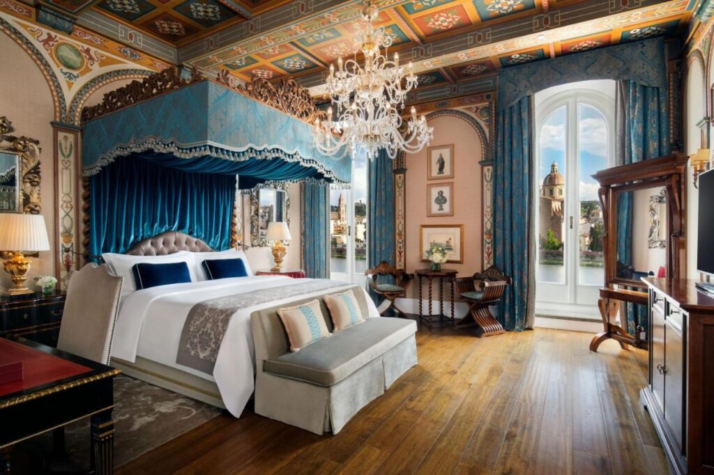 The 7 Best Luxury Hotels in Florence 3 Opulent bedroom at The St. Regis Florence with a grand blue canopy bed, crystal chandelier, detailed fresco ceiling, and arched windows framing Florence’s skyline.