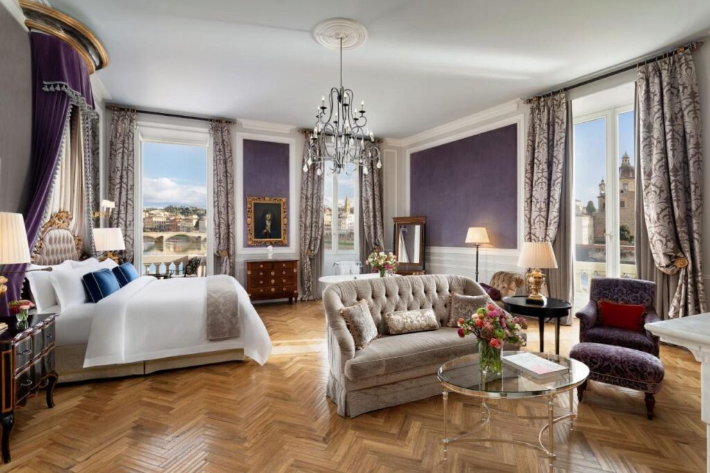 The 7 Best Luxury Hotels in Florence 4 Lavish suite at The St. Regis Florence featuring a regal bed with purple drapes, a plush beige sofa, glass coffee table, and panoramic views of the Ponte Vecchio.
