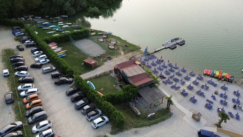 Aerial view of Bahia Cafè at Lago di Bilancino in Tuscany at the end of the day, showing empty loungers and umbrellas on the beach, colorful kayaks neatly arranged, and a crowded parking area as visitors gather for lakeside happy hour