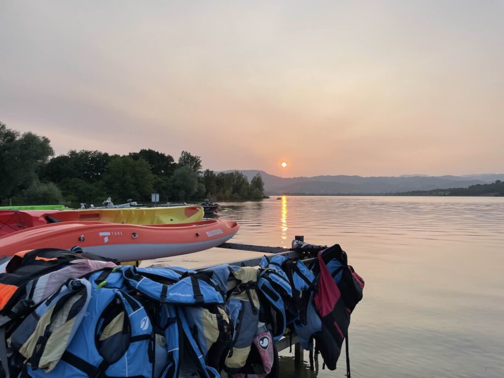 View of Lago di Bilancino at sunset with orange and yellow kayaks docked and life jackets hanging in the foreground, reflecting a peaceful summer atmosphere in Tuscany.