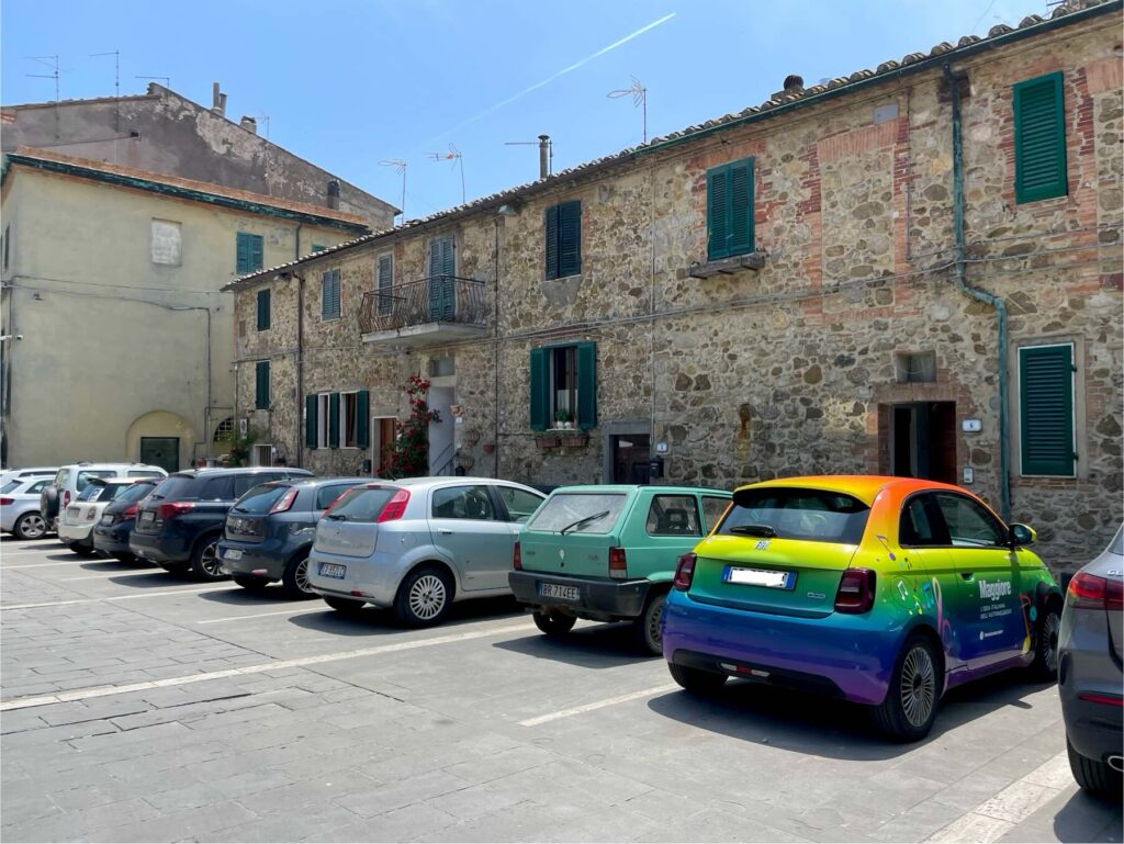 Brightly colored electric Fiat 500 parked in front of stone buildings in the historic center of Chiusdino, Tuscany.