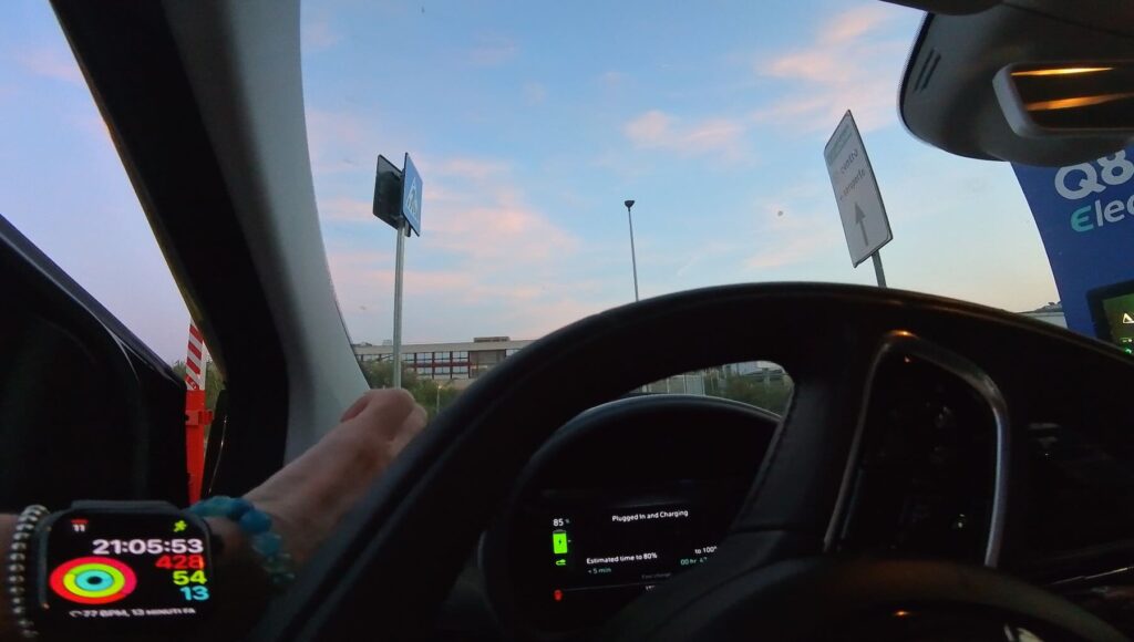 View from inside an electric car while charging at a Q8 station near Florence Airport, dashboard showing battery at 85% and estimated time to full charge.