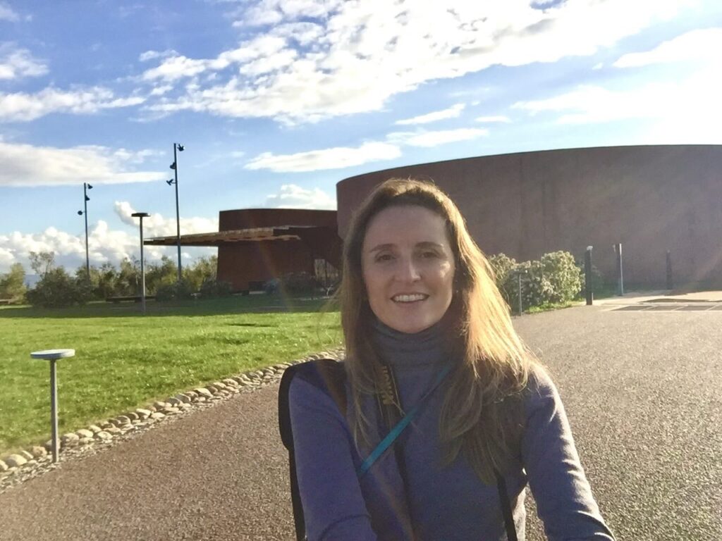 Woman smiling in front of Forum Bertarelli on a bright day, with green lawn, blue sky, and modern architecture in the background.