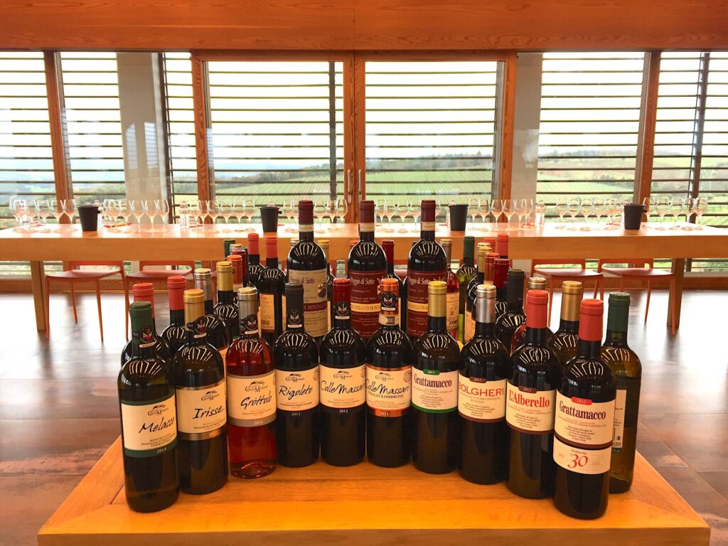 Bottles of ColleMassari, Grattamacco, and Poggio di Sotto wines arranged in a tasting room with large windows overlooking Tuscan vineyards.