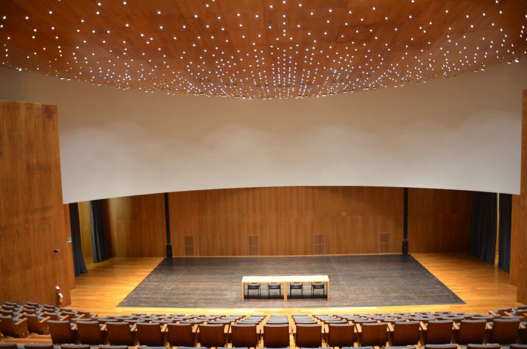 Empty auditorium of Forum Bertarelli with wooden walls, curved stage, and ceiling lights resembling stars.