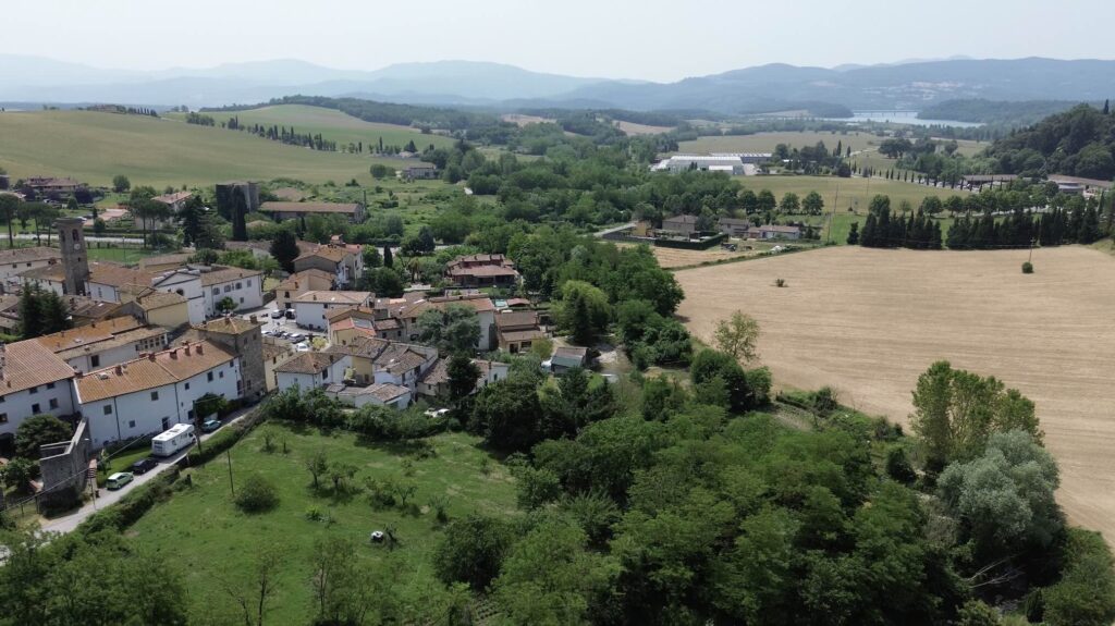 Aerial photo of a small village in the Mugello region of Tuscany, surrounded by green fields and hills, with Lake Barberino seen in the distance under a hazy summer sky.