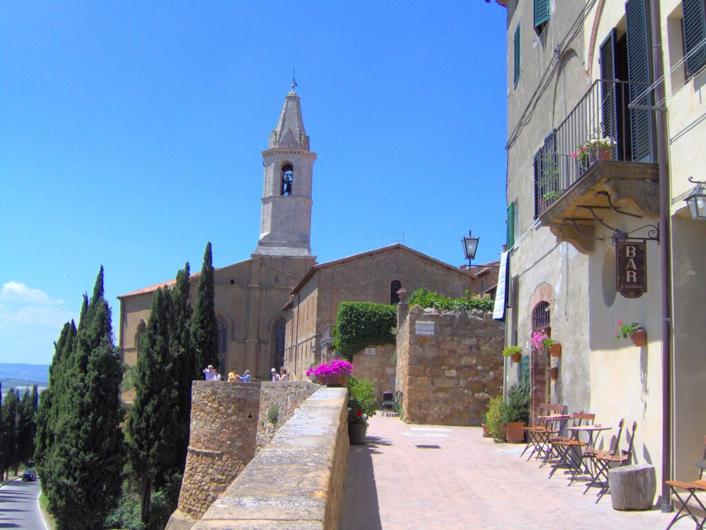 View of a sunny street in Pienza, Tuscany, with cypress trees, stone buildings, a bell tower, and outdoor café tables, capturing the peaceful charm of the Val d’Orcia region.