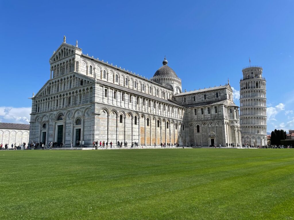 Wide-angle view of Pisa’s Cathedral and the Leaning Tower on a sunny day, with a green lawn and blue sky in the Piazza dei Miracoli, Tuscany, Italy.