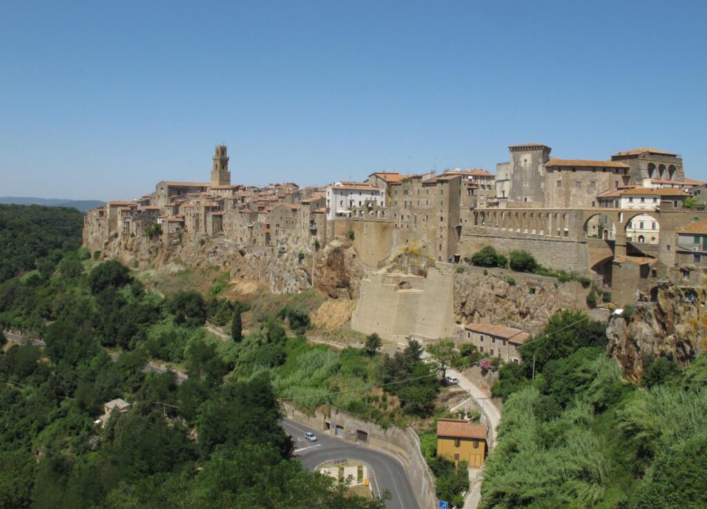 Panoramic view of Pitigliano, a medieval town in Tuscany built on a rocky cliff, with stone houses, towers, and walls rising above a green valley under a clear blue sky.