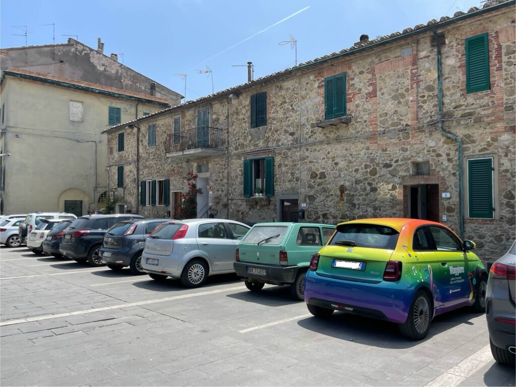 A brightly colored electric Fiat 500 parked in a small Tuscan village square, surrounded by stone buildings and other cars—ideal example of flexibility when renting a car in Florence.