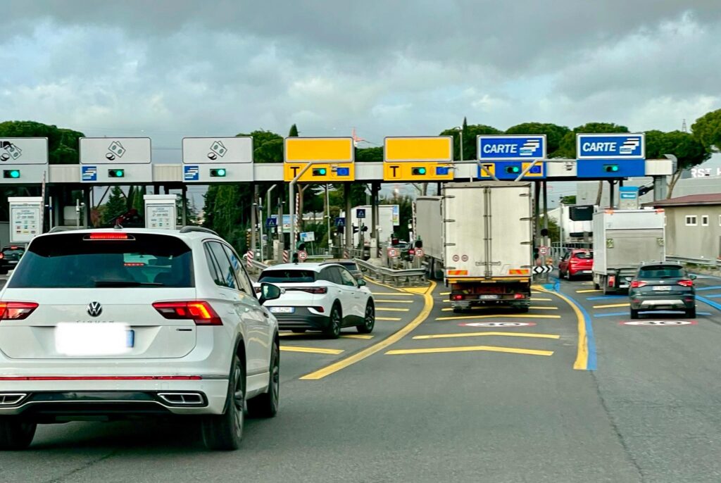 Cars approaching toll booths on an Italian autostrada, with yellow Telepass-only lanes and blue lanes for card payments. A Volkswagen SUV is visible in the foreground under cloudy skies.