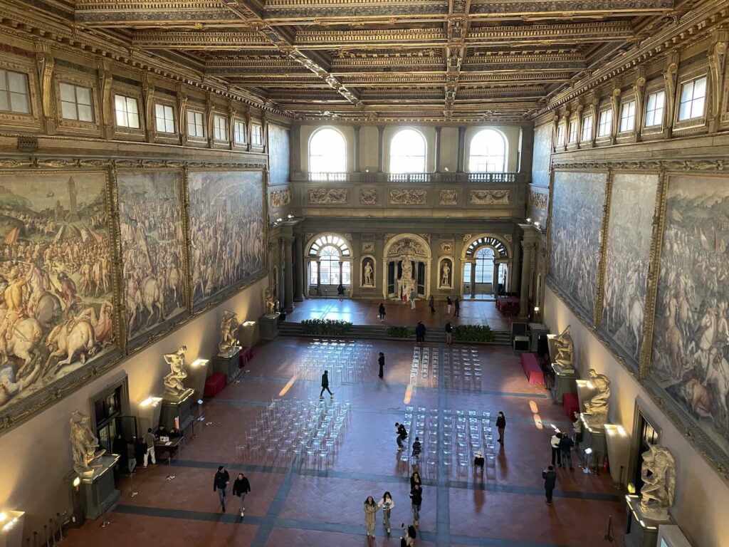 Salone dei Cinquecento inside Palazzo Vecchio in Florence with large Renaissance frescoes, statues, and seating arranged for a ceremony.
