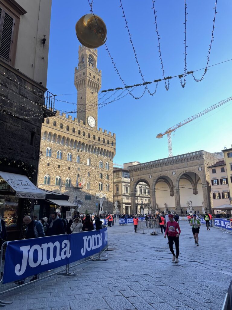 Runners participating in the 2023 Florence Marathon cross Piazza della Signoria on a sunny November day, with Palazzo Vecchio and early Christmas decorations visible under a clear blue sky.