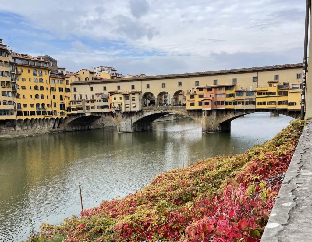 View of Ponte Vecchio over the Arno River in Florence on an October day, with red and orange autumn leaves in the foreground.