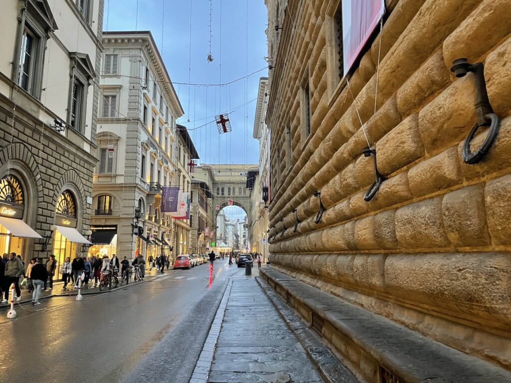 View of Via degli Strozzi in Florence on a rainy November day, showing the rusticated façade of Palazzo Strozzi and the illuminated arch of Piazza della Repubblica decorated for the holiday season.