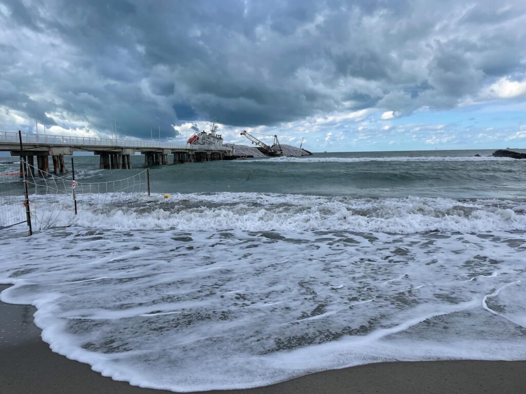 Photo of the Guang Rong cargo ship stranded next to the pier in Marina di Massa, Tuscany, with rough sea waves and a cloudy sky in the background.