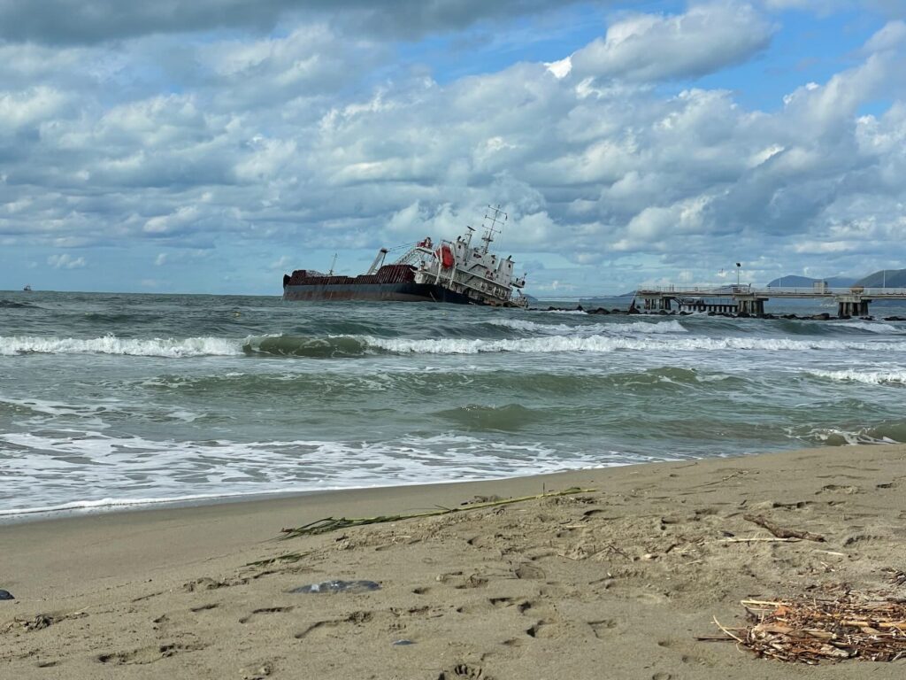 View from the beach of Marina di Massa showing the Guang Rong cargo ship stranded near the pier, with waves rolling in and a cloudy sky above.