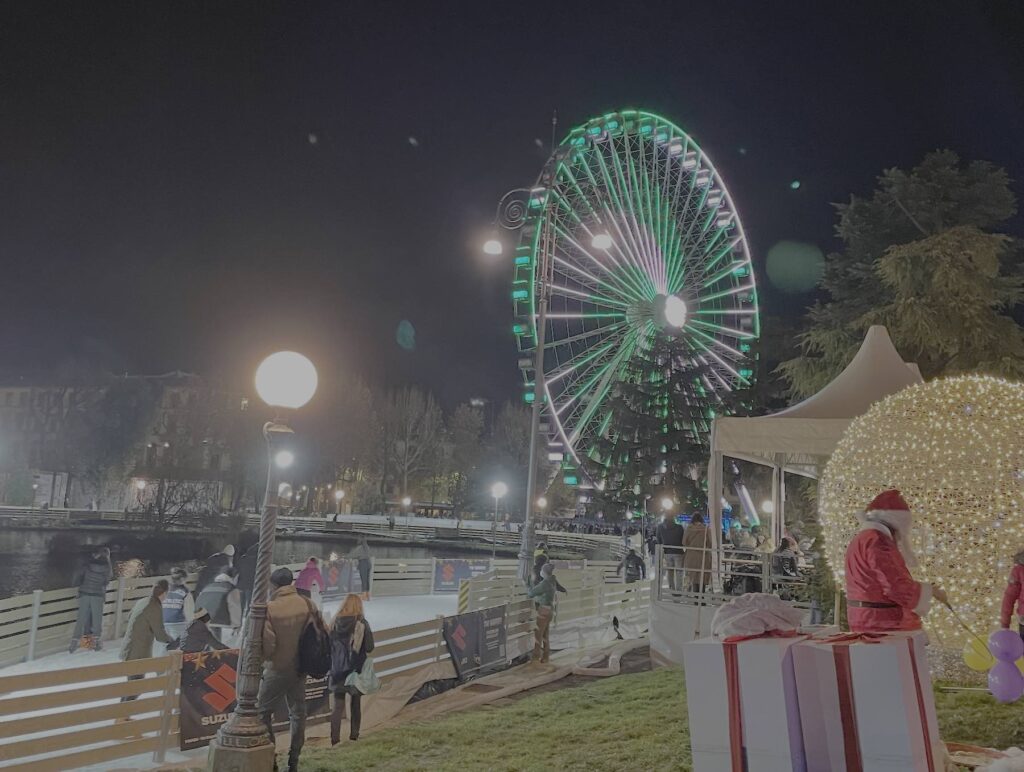 Night view of the Florence Eye Ferris wheel illuminated in green at Fortezza da Basso, with people ice-skating nearby and Christmas decorations creating a festive winter atmosphere.