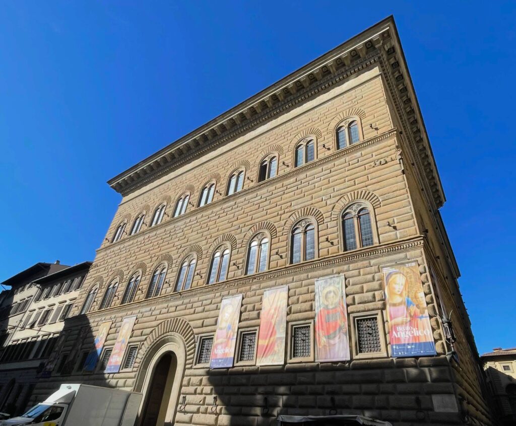 Exterior of Palazzo Strozzi in Florence with large banners promoting the Beato Angelico exhibition, seen under a bright blue sky.