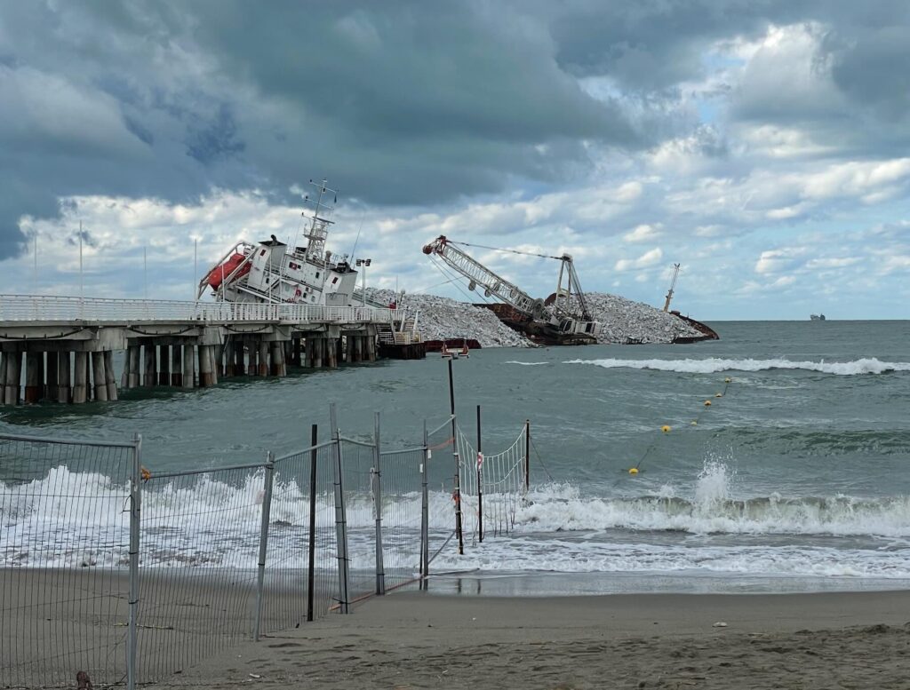 Guang Rong cargo ship stranded near the Marina di Massa pier, with construction equipment and stone cargo visible, photographed on September 26, 2025 under cloudy skies.