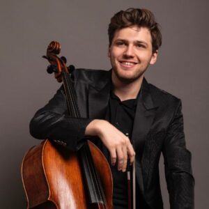 Christmas Concert in Florence 2025: Classical Music at Teatro Verdi on Christmas Eve 2 Portrait of a young classical cellist holding a cello, wearing a dark jacket and smiling, photographed against a neutral background.