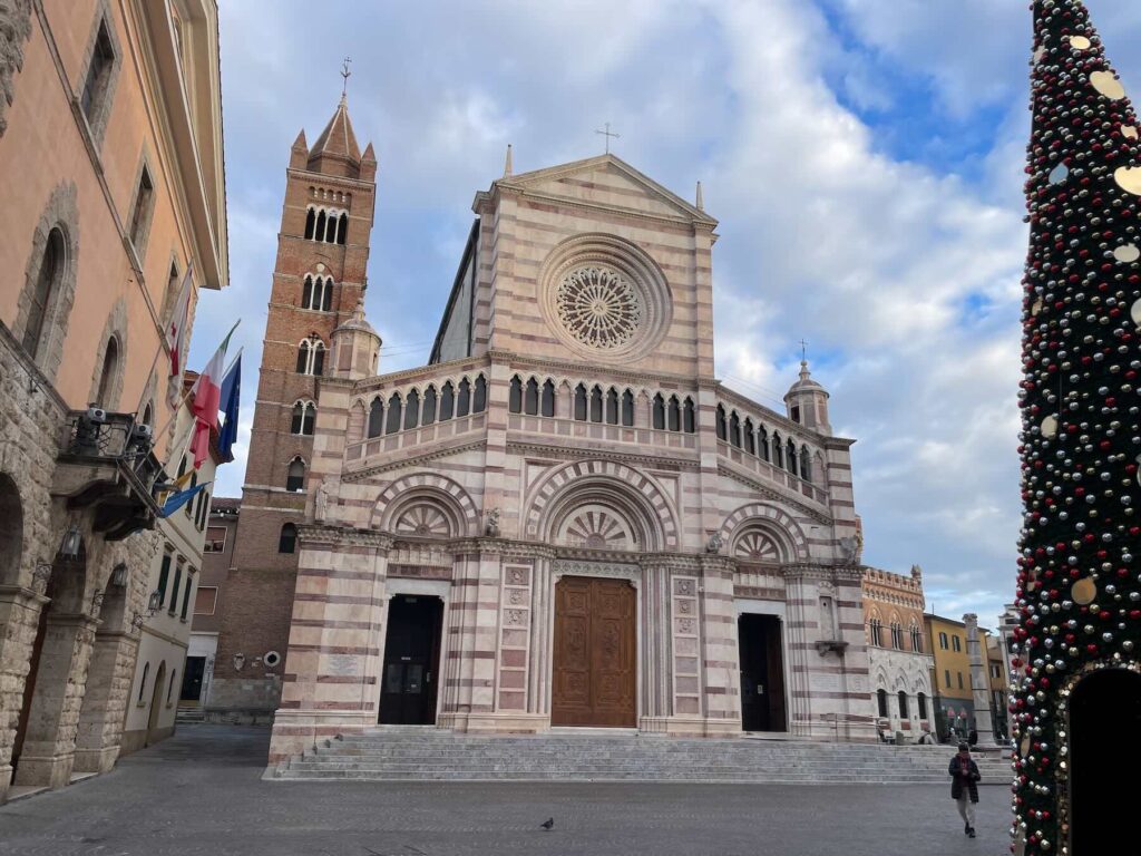 Front view of the Cattedrale di San Lorenzo in Grosseto, showing its striped marble façade and bell tower, with a decorated Christmas tree on the side.