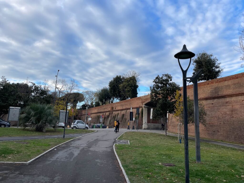 View of the Medici Walls at the entrance to Grosseto’s historic center, with a walkway, street lamps, trees and a cloudy winter sky.