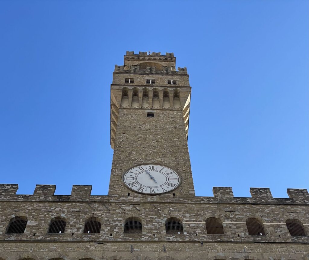 Upward view of Palazzo Vecchio’s Arnolfo Tower with its clock and stone façade, photographed on a clear day; the site is among the Florence Civic Museums introducing new ticket prices from February 1, 2026