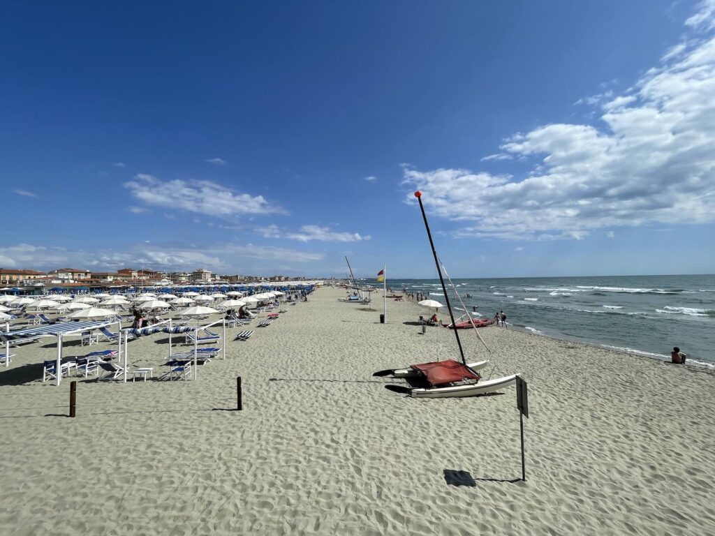 La Prima Estate 2026: the Tuscany festival that feels like a beach vacation 14 : Wide sandy beach in Lido di Camaiore, Versilia (Tuscany), with rows of white umbrellas and lounge chairs on the left and the Tyrrhenian Sea on the right under a bright blue sky.