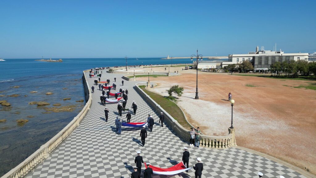 Participants in a ceremonial parade walk along Livorno’s seafront during Settimana Velica carrying national flags