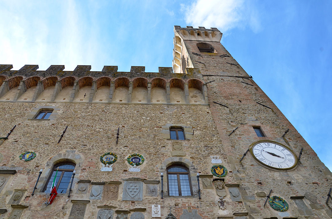 Low-angle view of the Palazzo dei Vicari in Scarperia showing the stone façade, clock, tower, and decorative coats of arms.