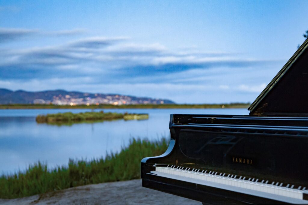 Free Classical Music Concerts in Orbetello 2026: Young Pianists Compete on the Tuscan Coast 1 Grand piano by the Orbetello lagoon in Tuscany, with water, reeds, and the town in the background.