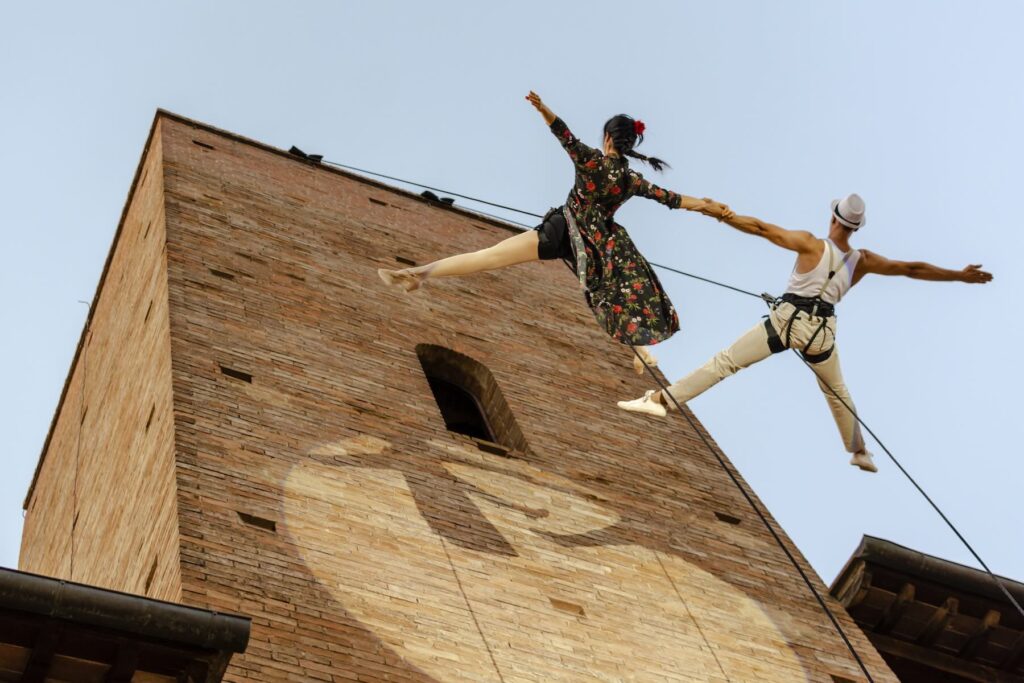 Aerial performers holding hands above a brick building during Mercantia street theater festival in Certaldo, Tuscany.
