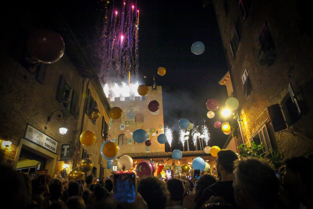 Night scene at Mercantia in Certaldo with colorful balloons, lights, and a crowd in front of medieval buildings.
